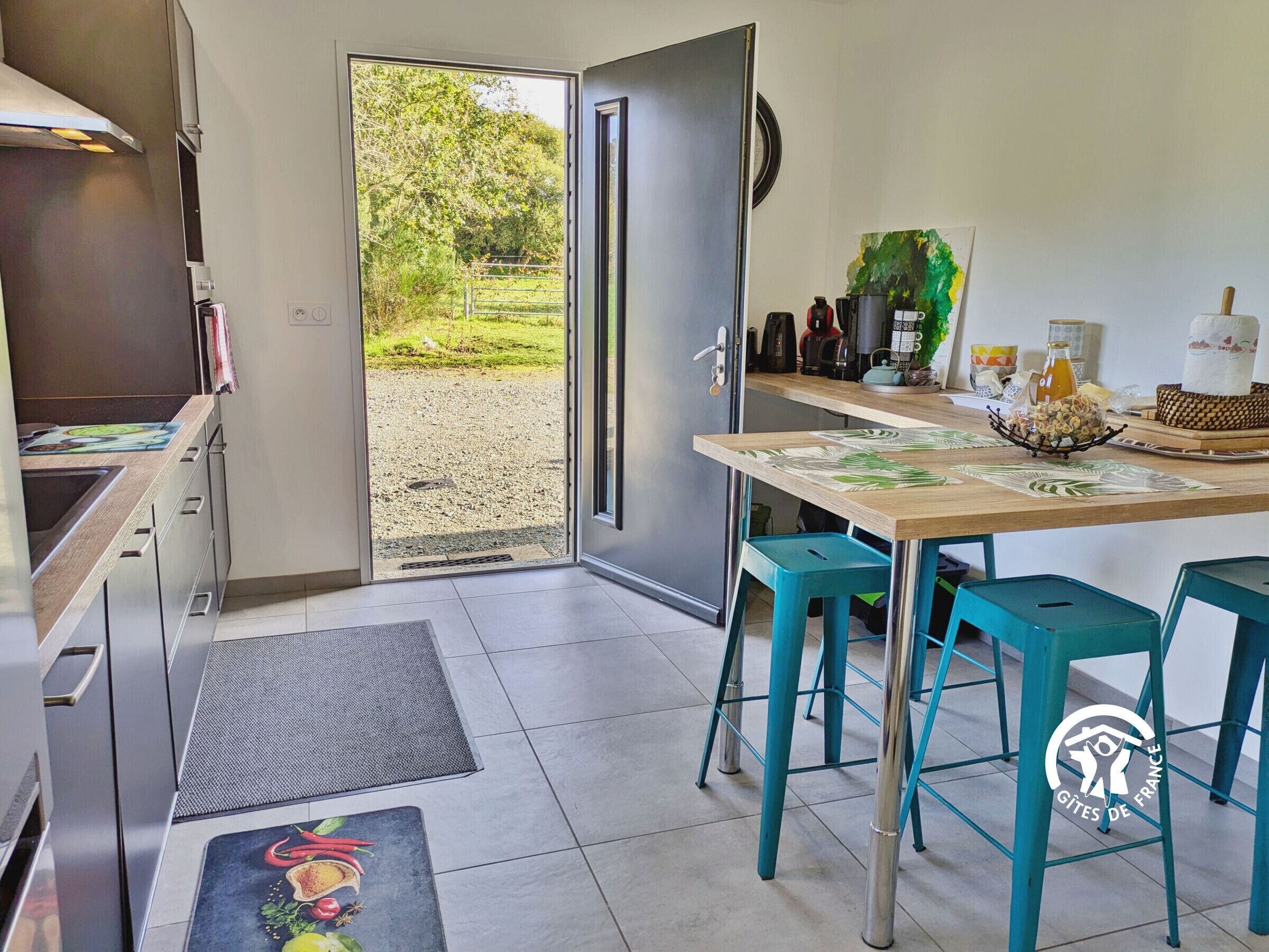 A kitchen with a view of a garden, a dining area with stools, and a kitchen counter with appliances.