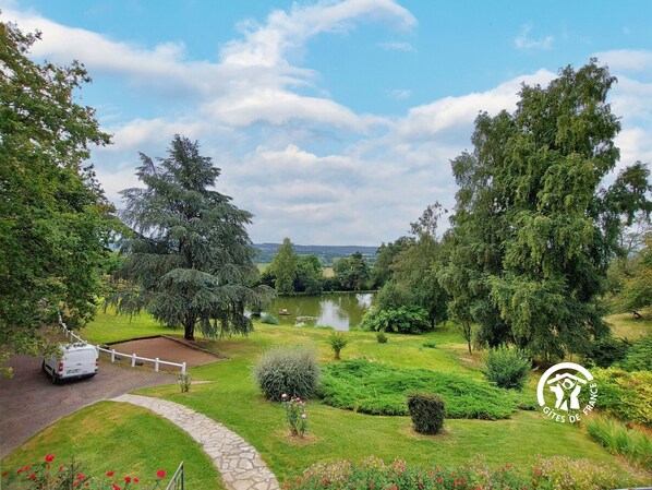 Un jardin paisible avec un étang, une végétation luxuriante et un fourgon blanc garé près d’un sentier.