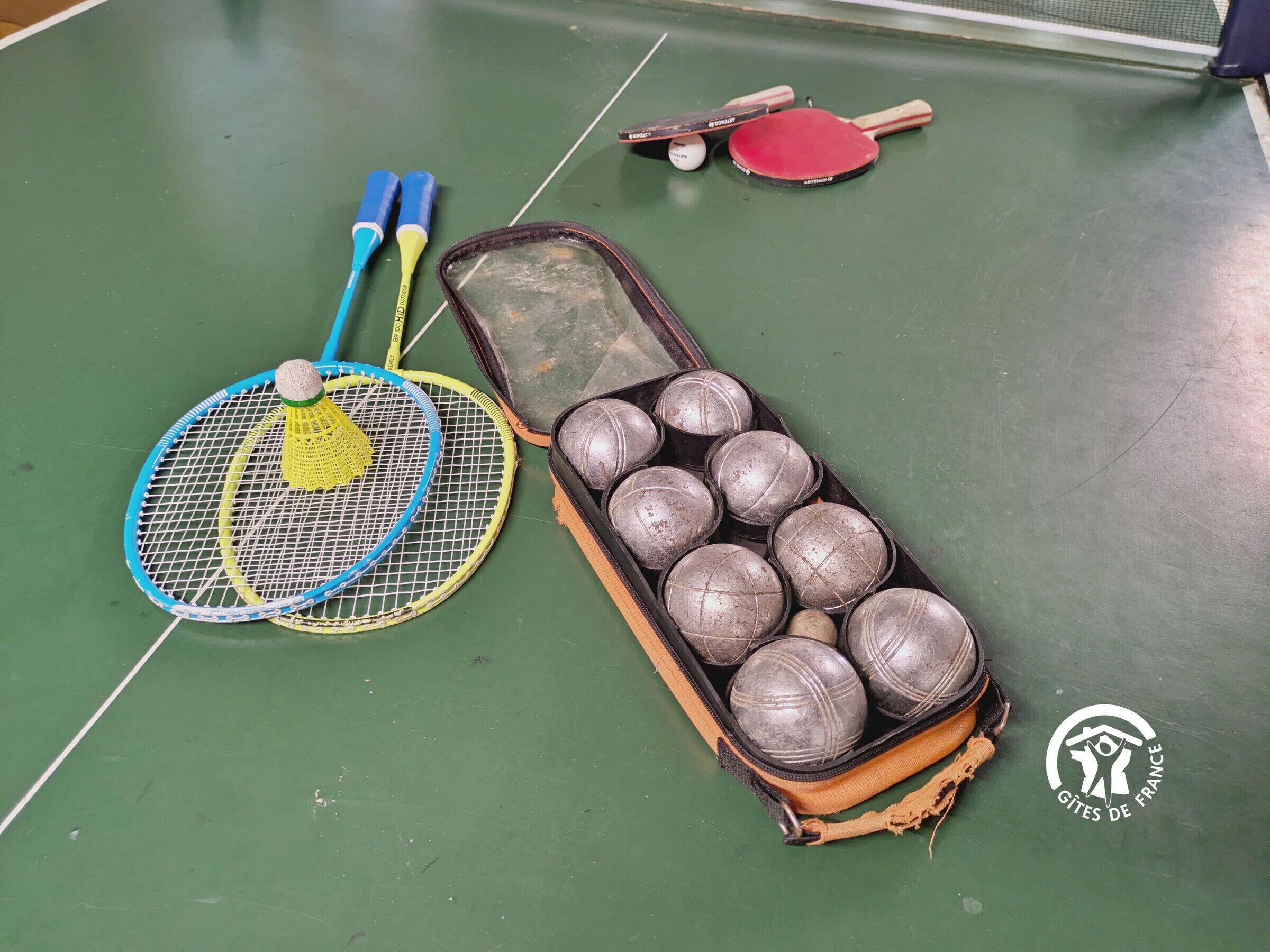 A table with a blue and yellow table tennis paddle, a red paddle, and a case containing multiple table tennis balls.
