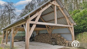 Una estructura de madera con techo inclinado, suelo de hormigón y una pila de leña al fondo.