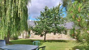 A patio with a table and chairs, a weeping willow tree, a small tree, and a stone building in the background. - Cozy Countryside Home with Private Garden, Near Laval - 4 Bedrooms, Pet-Friendly, Ideal Location (Saint-Ouën-des-Toits)