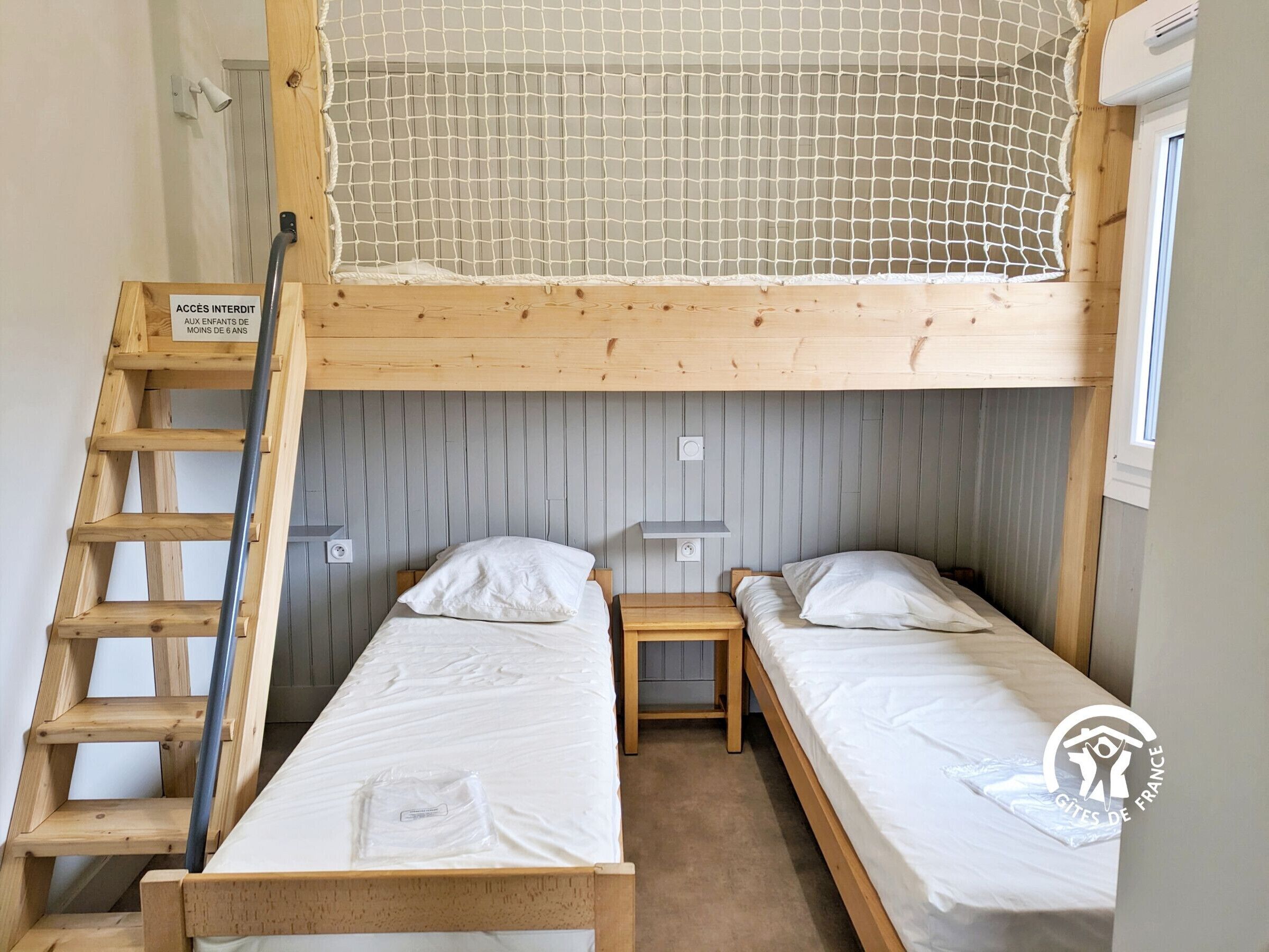 Quarto com beliche, escada, duas camas simples, uma mesa pequena e aviso proibindo a entrada de crianças menores de 6 anos.