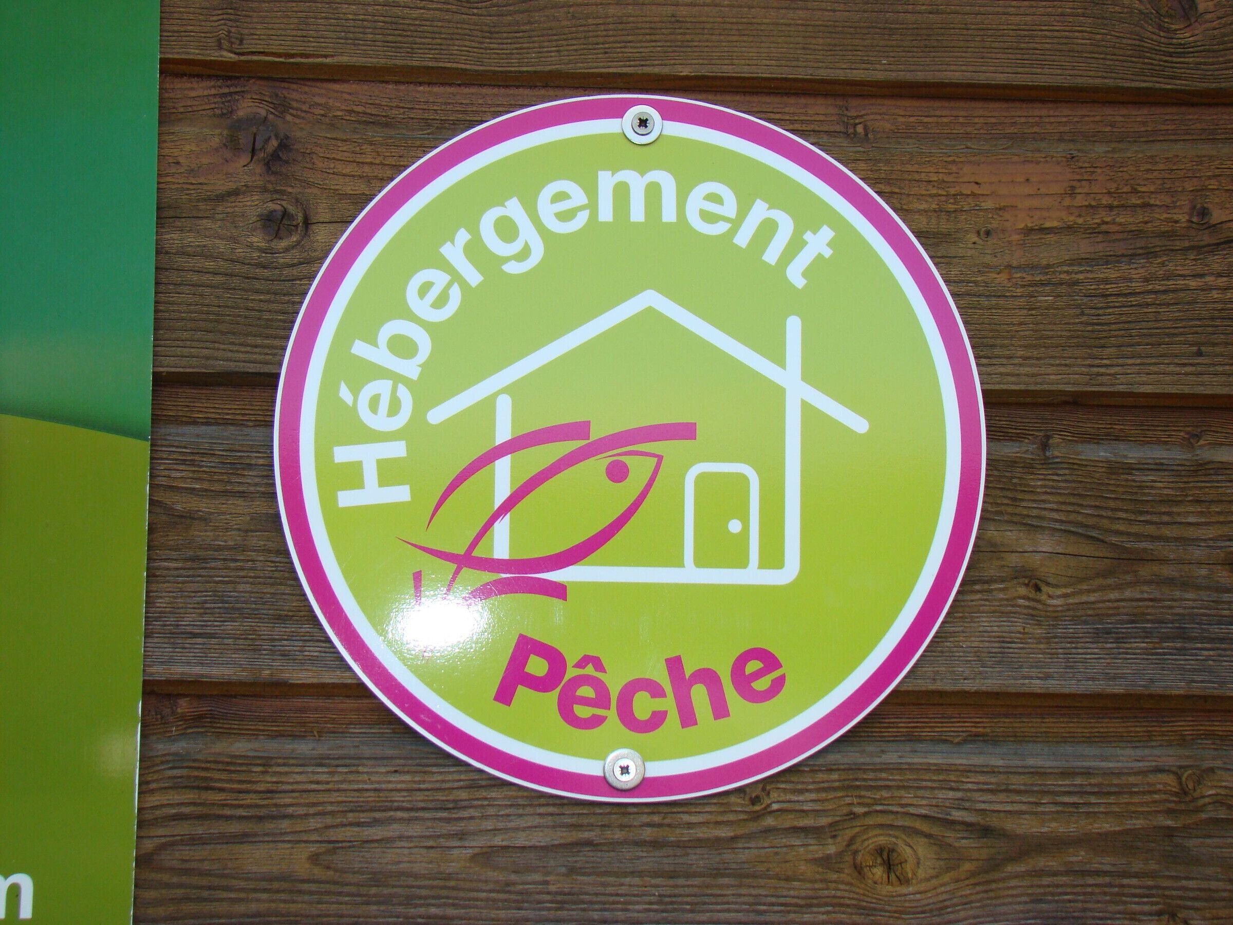 Ein rundes Schild mit dem Aufdruck "Hébergement Pêche" und einem Haus-Symbol, angebracht an einer Holzwand.