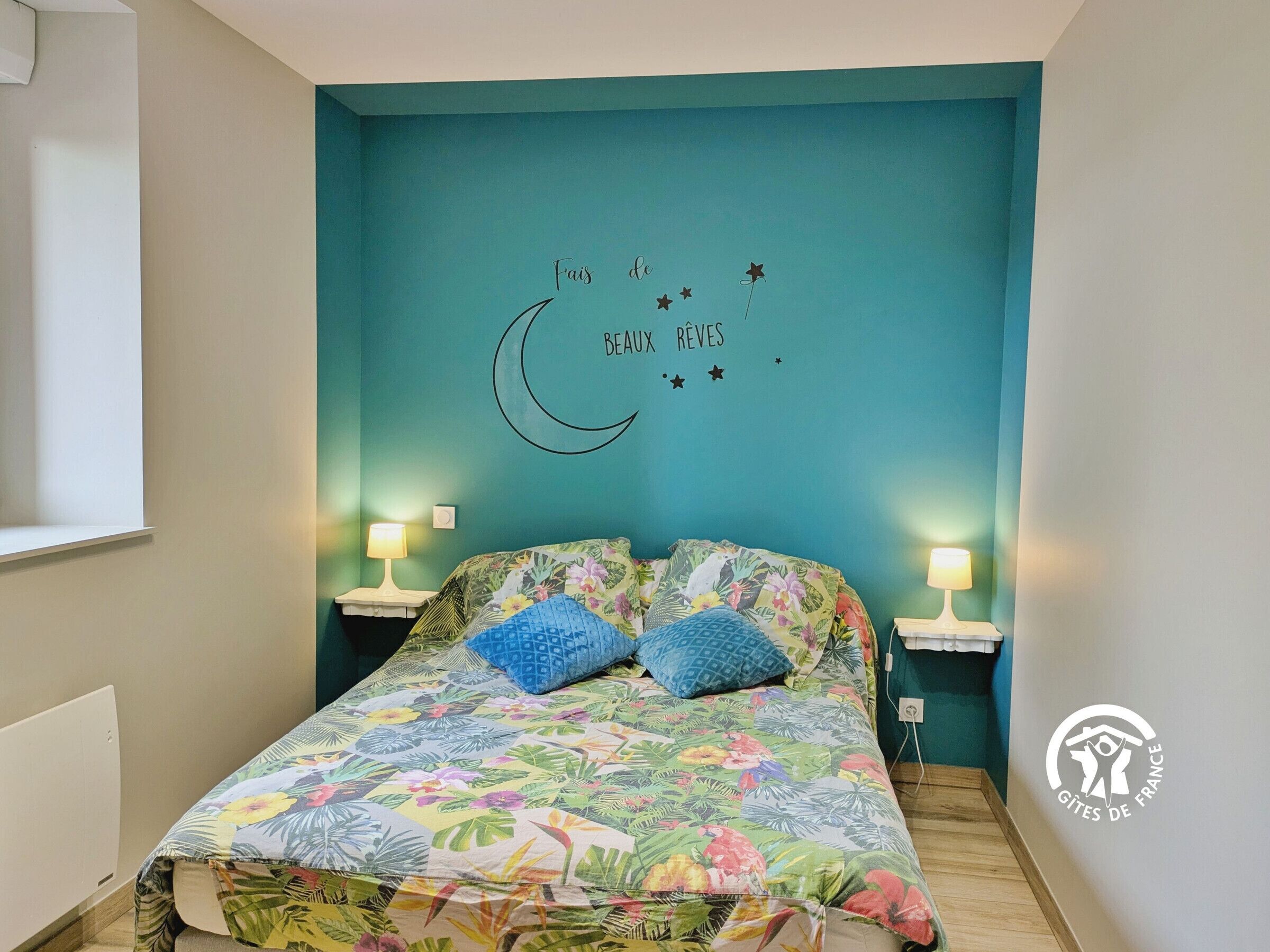 Ein Schlafzimmer mit einer türkisfarbenen Wand, einer blumigen Tagesdecke, zwei blauen Kissen und einem Nachttisch mit Lampe.