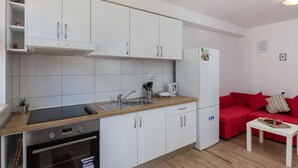 Cocina moderna con armarios blancos, horno empotrado, lavaplatos y refrigerador. En la sala de estar, hay un sofá rojo y una pequeña mesa blanca.