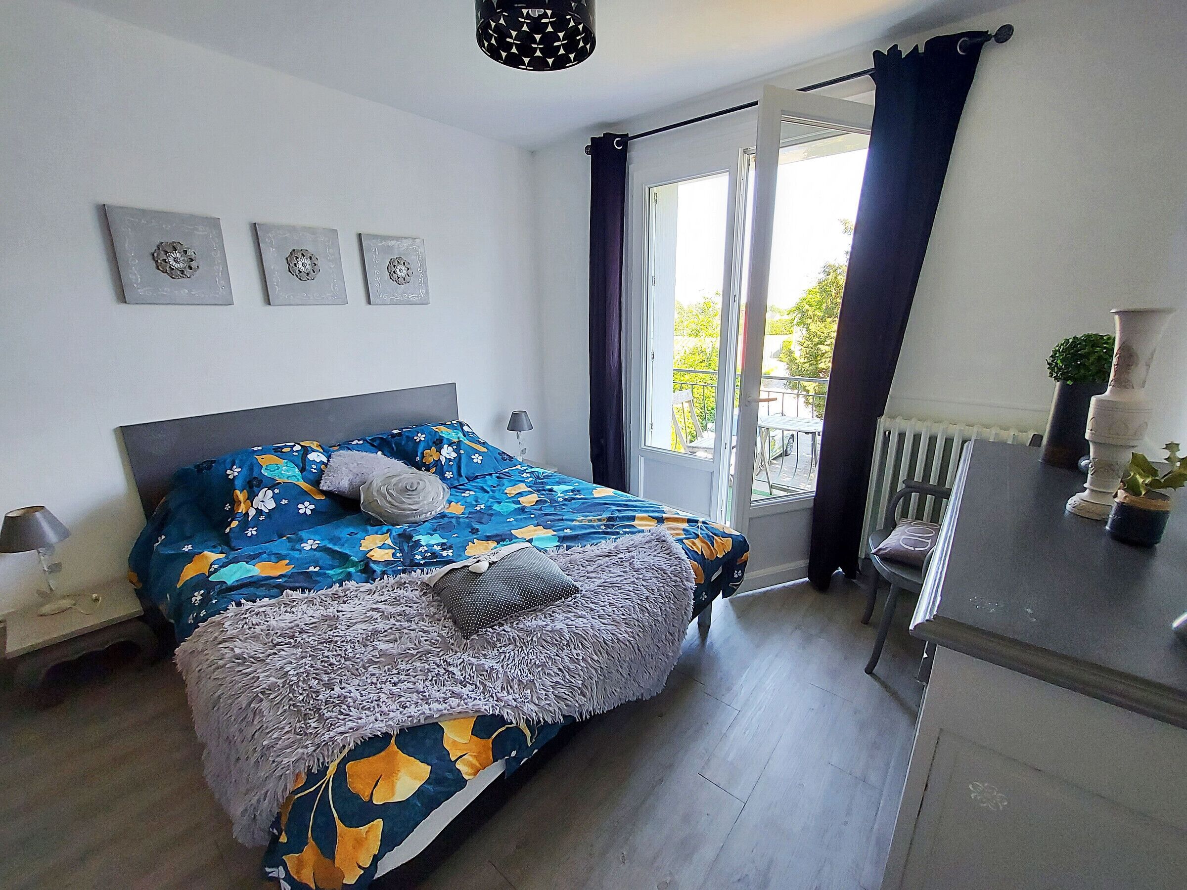A bedroom with a bed, a window with curtains, a radiator, and a small table with a plant.
