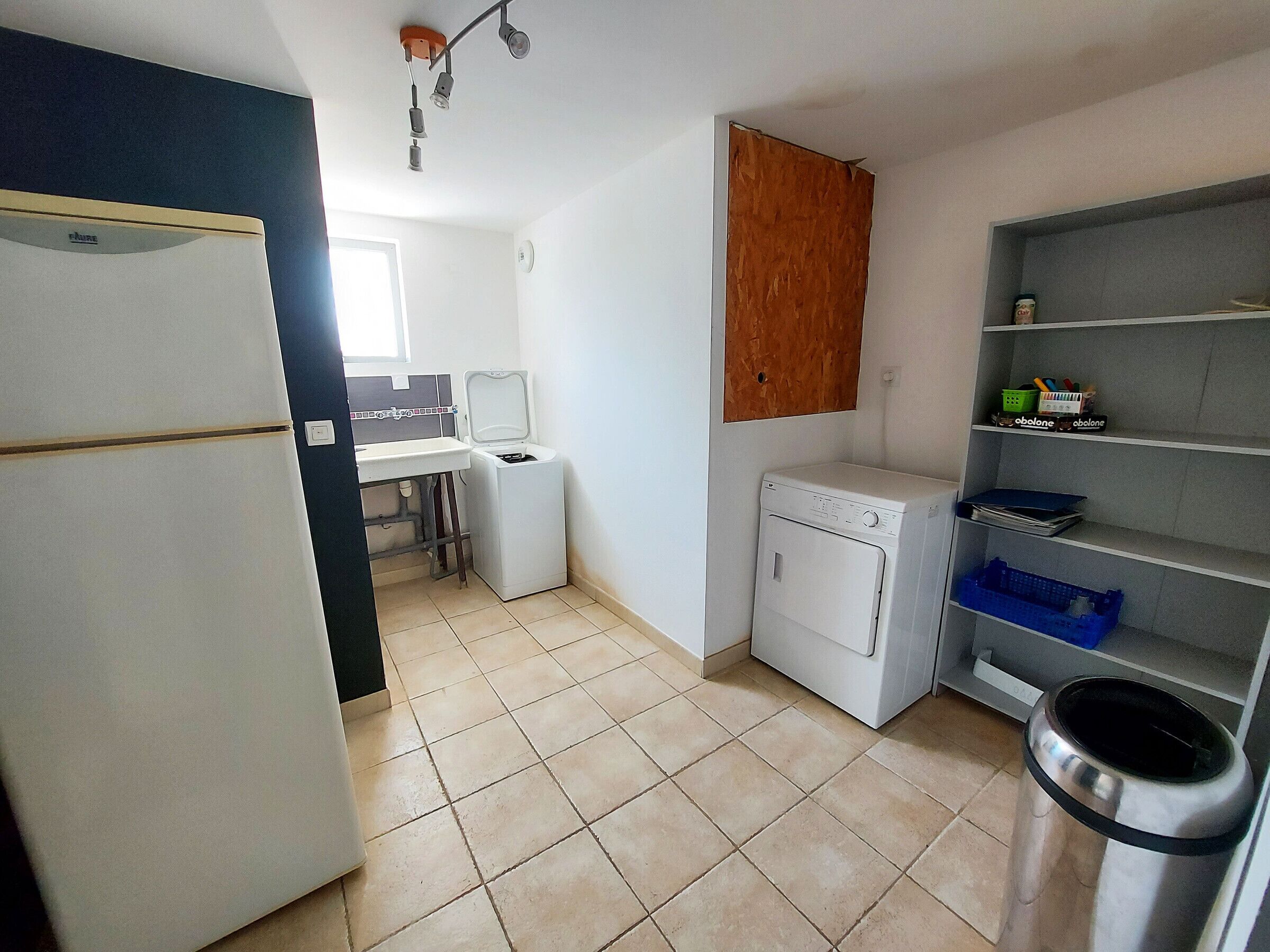 A compact kitchen with a refrigerator, washing machine, and a small sink. There is a shelf with storage bins and a trash can.
