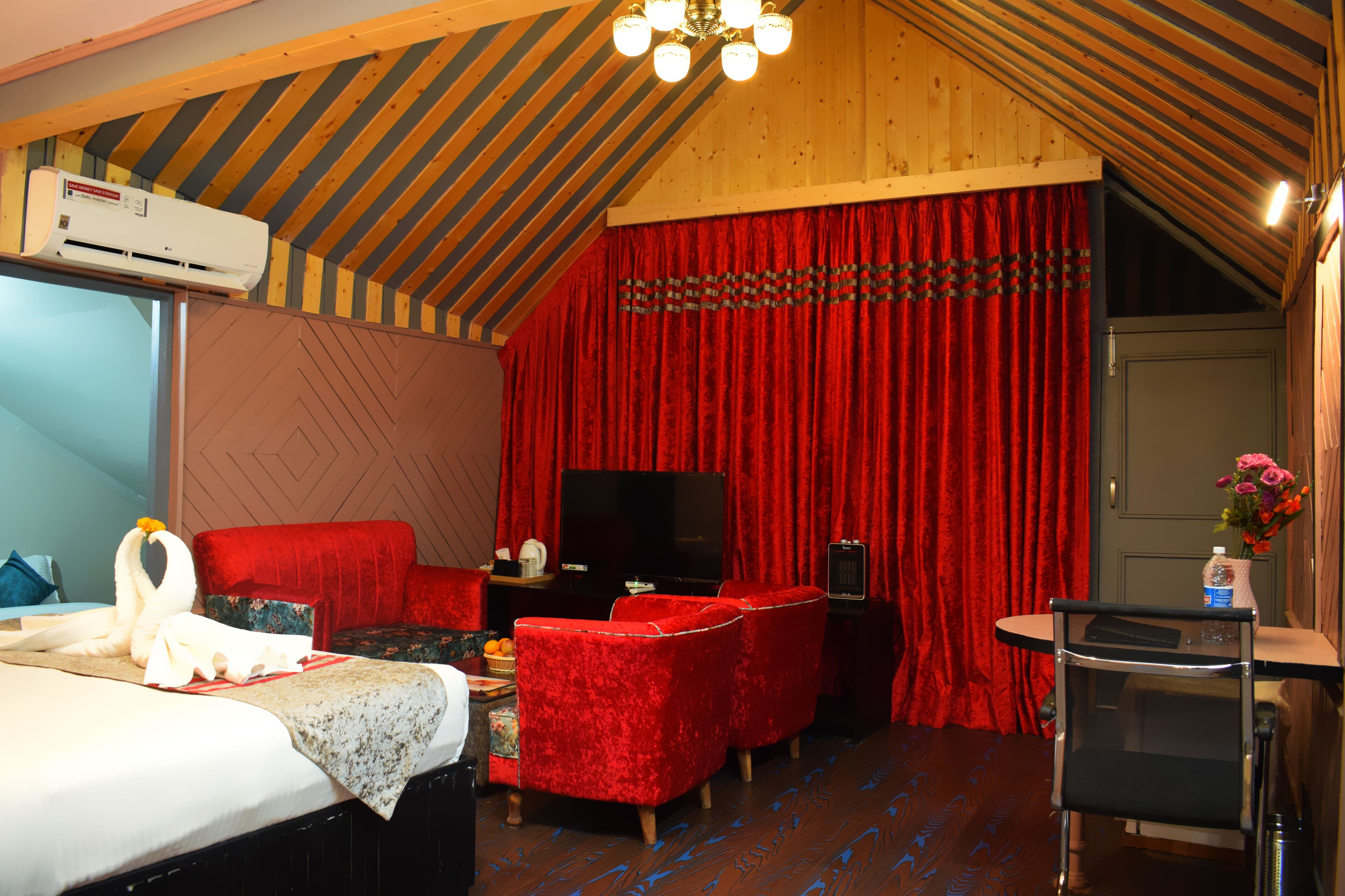 A hotel room with a bed, red velvet sofa, a flat-screen TV, and a small table with a vase of flowers.
