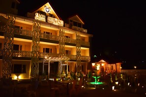 Un hotel de varios pisos con un cartel llamativo que dice “ONE HERITAGE KUD”. Cuenta con asientos exteriores con mesas y sillas, pilares decorativos y una pequeña fuente.
