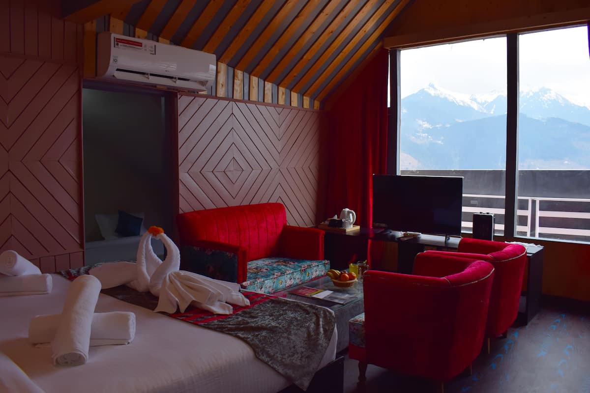 A hotel room with a bed, red chairs, a TV, and a mountain view.