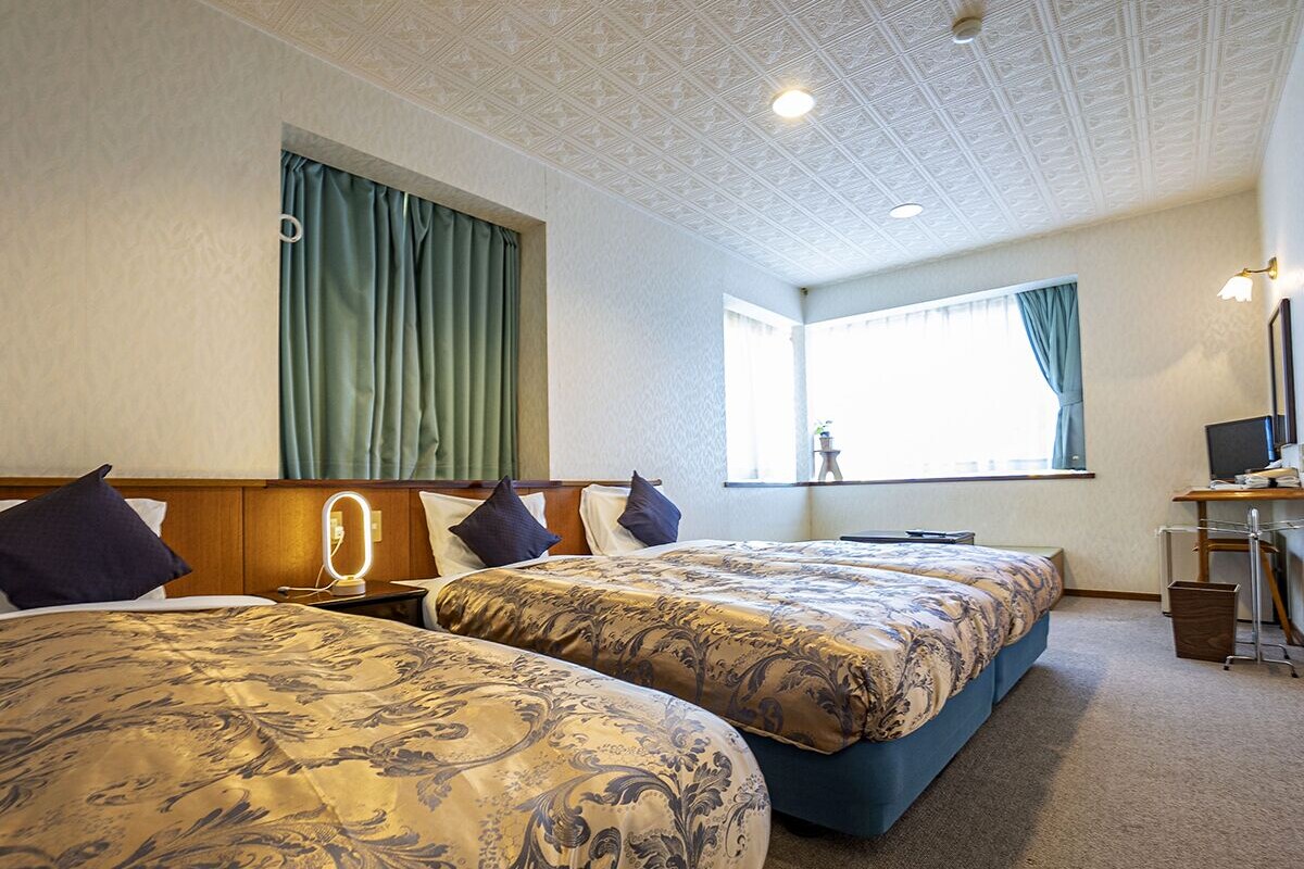 A hotel room with two beds, a desk, a chair, a lamp, and a window with curtains.