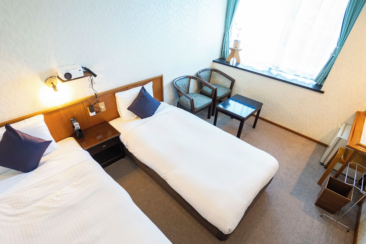 A hotel room with two beds, a desk, a chair, a window with curtains, and a bedside table with a lamp.