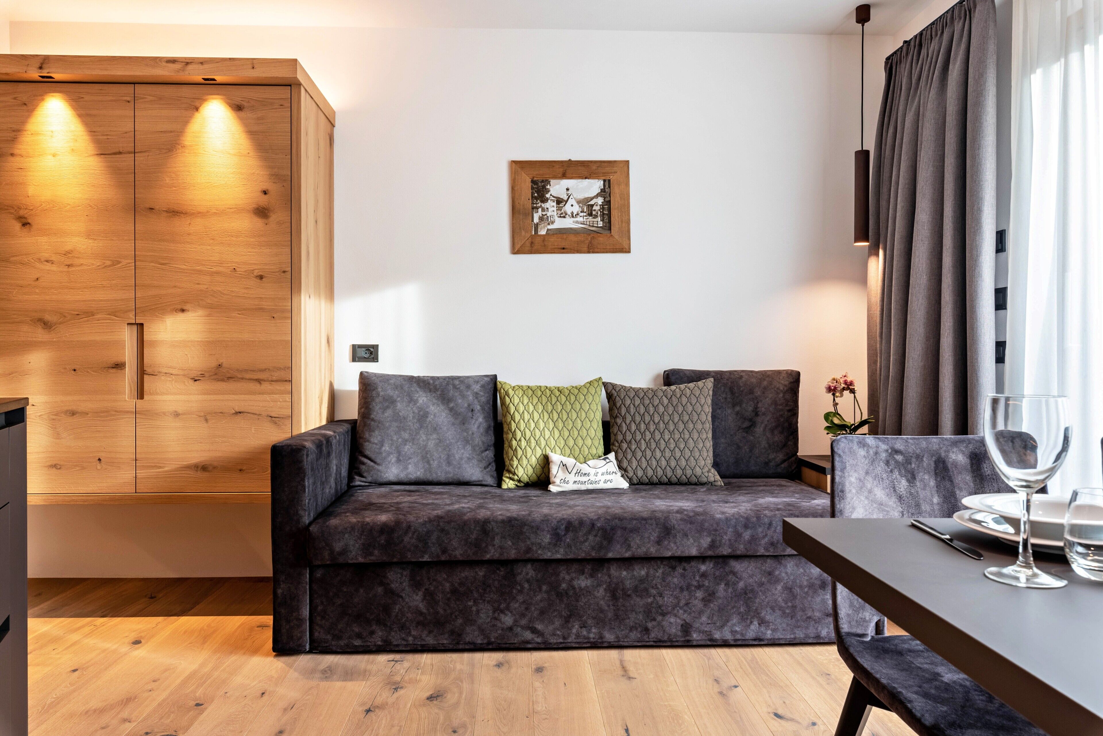 A modern living room with a dark grey sofa, wooden wardrobe, and a dining table with a wine glass.