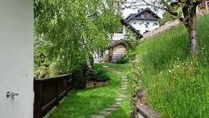 Ein schmaler Steinweg führt zu einer Holzhütte, umgeben von üppigem Grün und einem weißen Gebäude im Hintergrund.