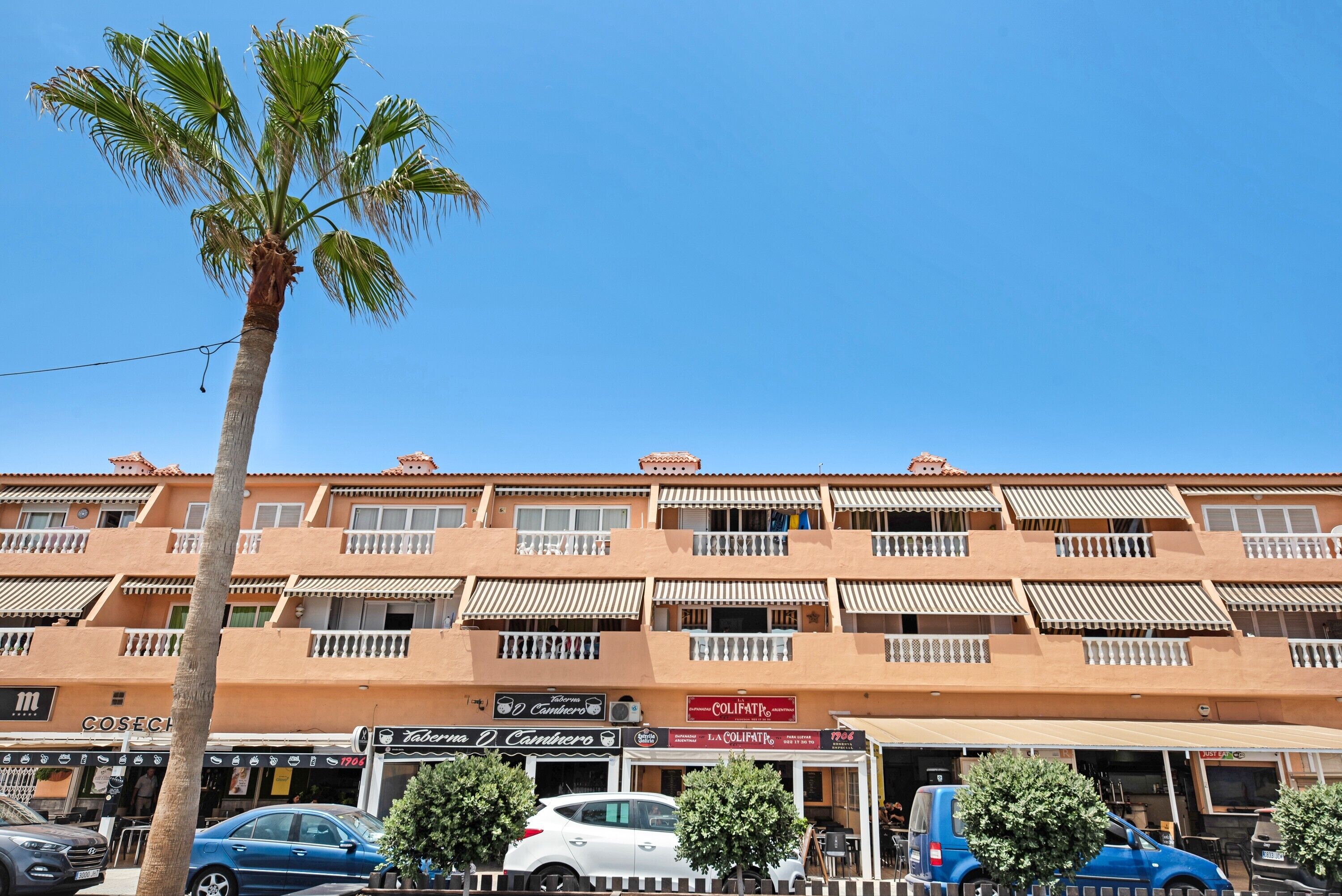 Un edificio de varios pisos con balcones, una palmera y varios coches estacionados al frente.