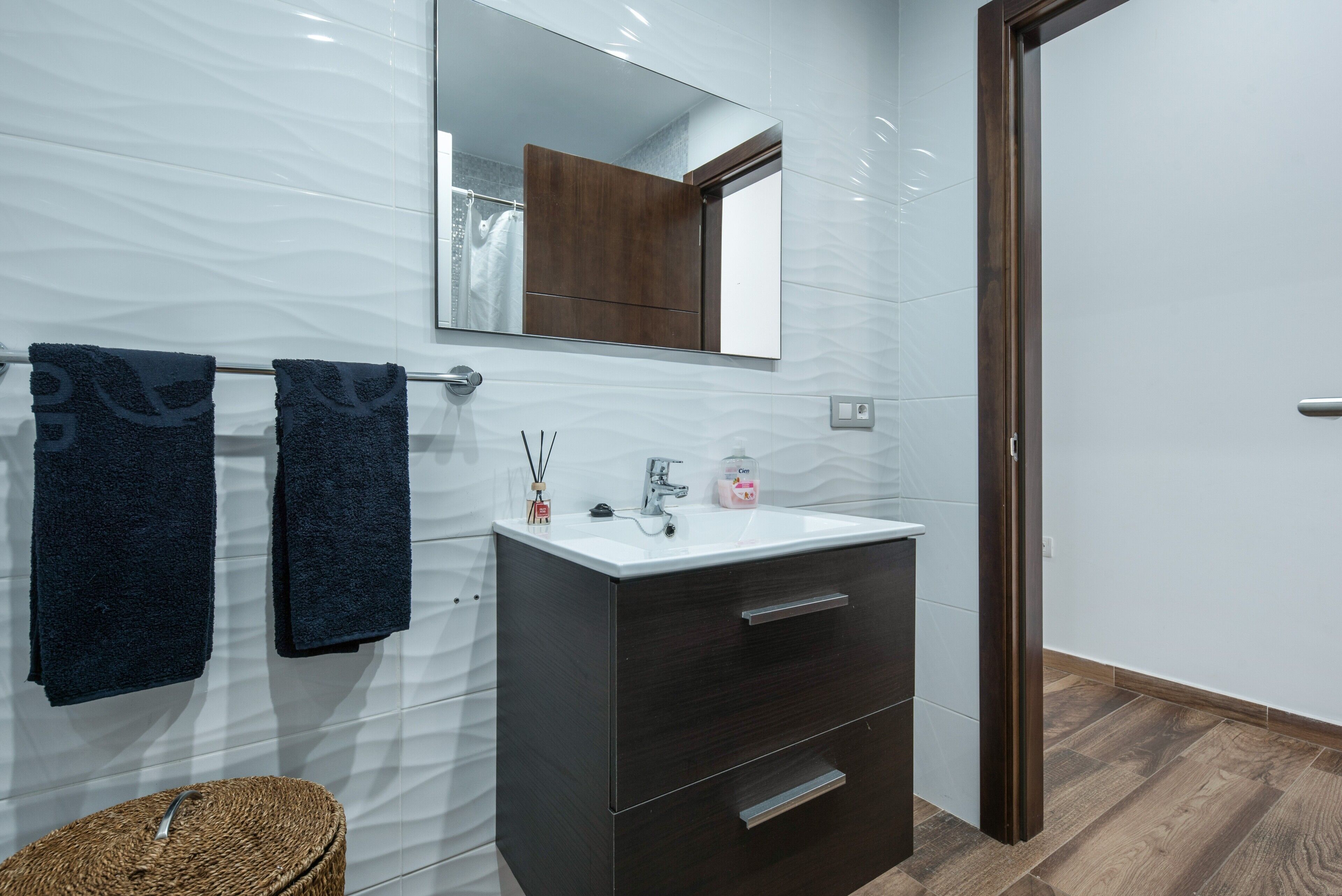 Une salle de bain avec un mur recouvert de carreaux blancs, un meuble en bois foncé, un lavabo blanc et deux serviettes accrochées au mur.