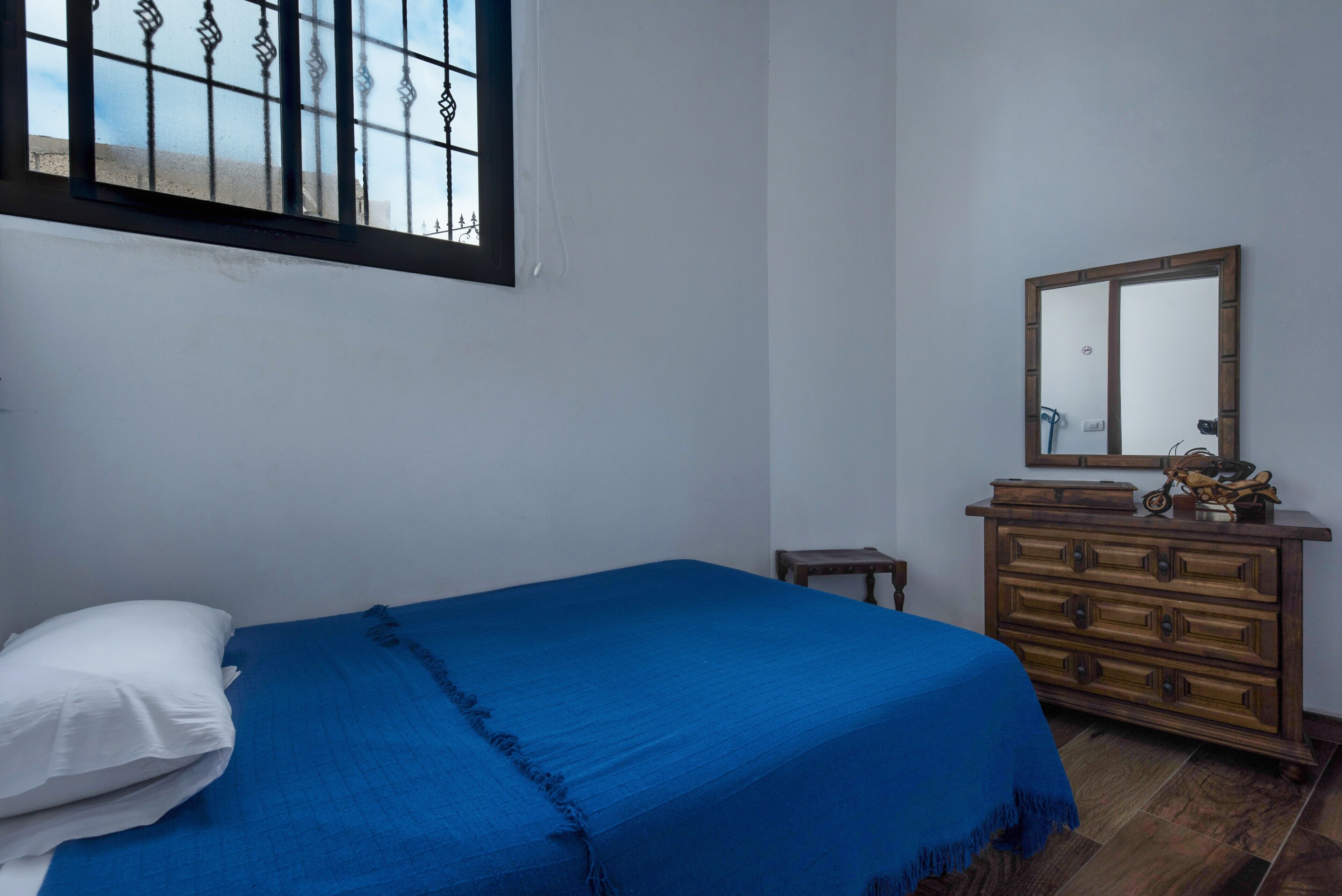 A bedroom with a bed, a dresser, a mirror, and a window with metal bars.