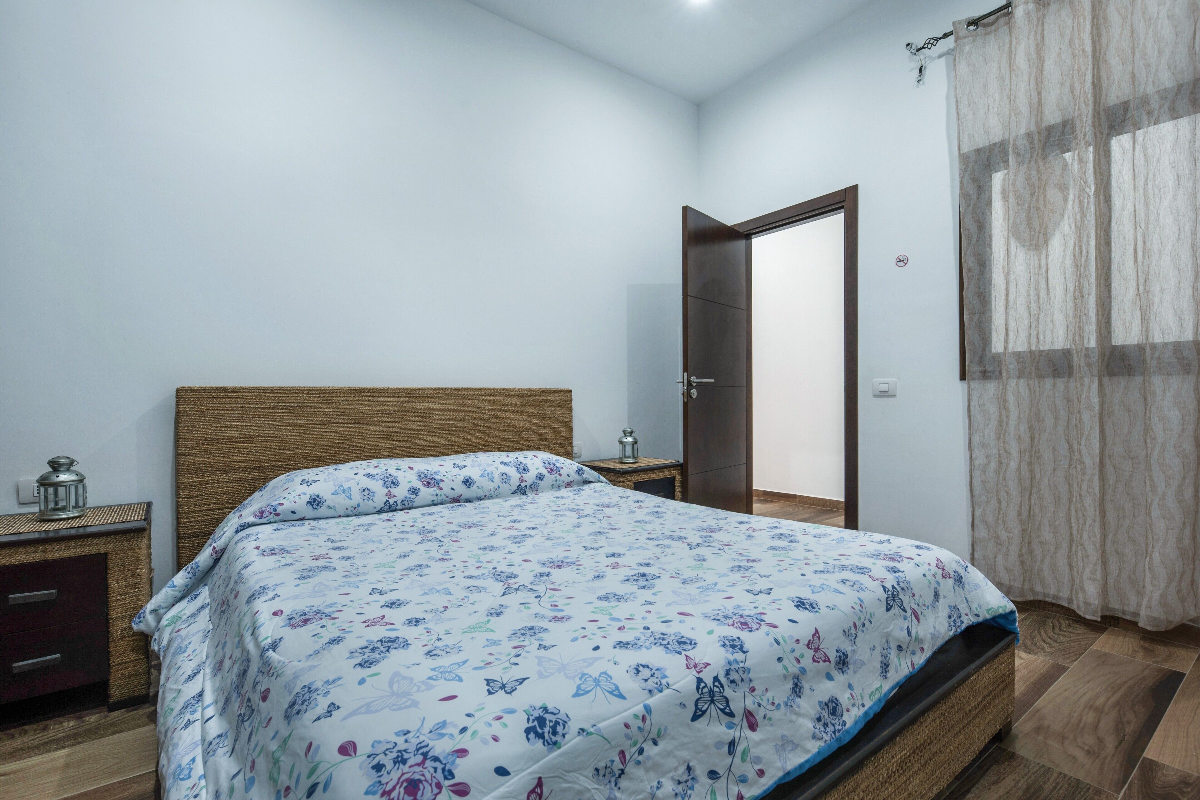 Ein Schlafzimmer mit Bett, Nachttisch, Spiegel, Tür und einem Fenster mit Vorhängen.