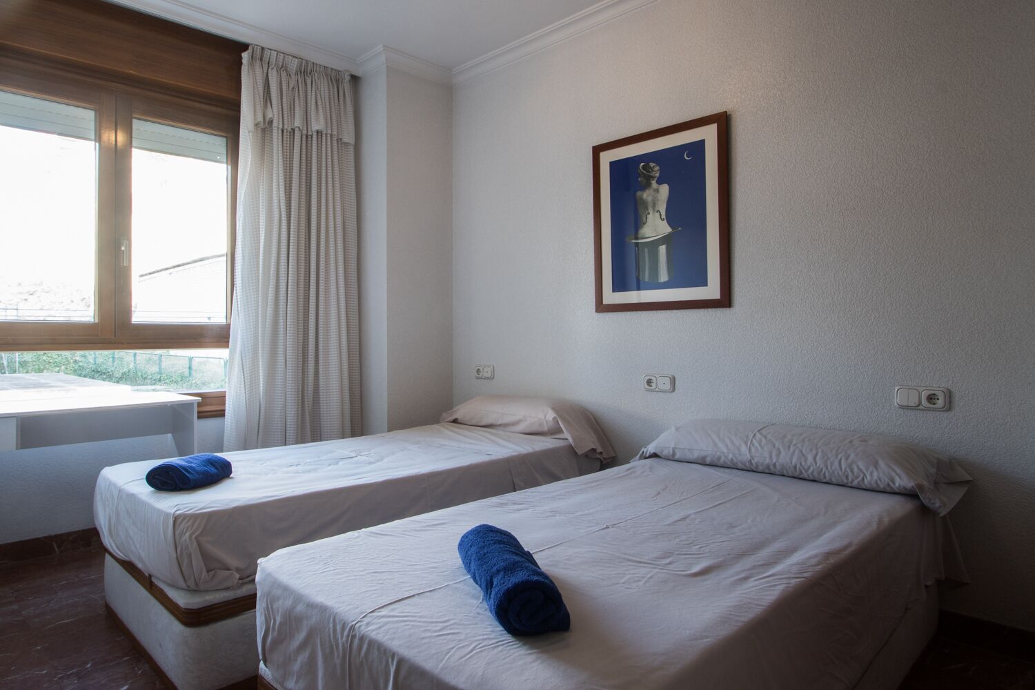 Una habitación con dos camas individuales, una ventana con cortinas, una mesita y un cuadro en la pared.
