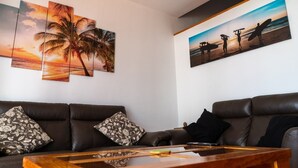 Ein Wohnzimmer mit einem braunen Ledersofa, einem Holztisch und zwei an der Wand hängenden Bildern mit tropischen und Strandmotiven.