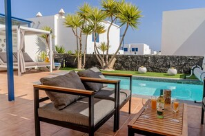 A poolside area with a sofa, a table with drinks, and a view of the pool and buildings.