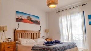 Ein Schlafzimmer mit einem Holzbett, zwei Nachttischen, einer Wandlampe, einem großen Fenster mit durchsichtigen Vorhängen und einem Gemälde einer Wüstenlandschaft.