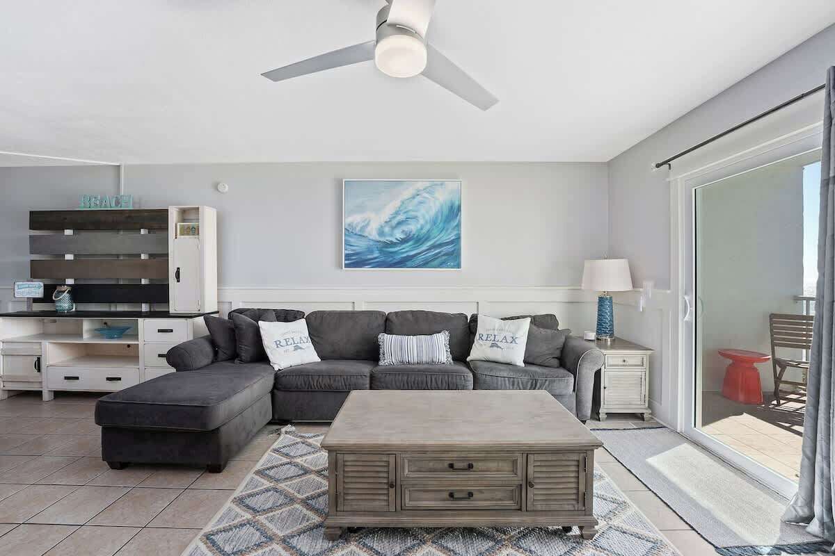 A modern living room with a grey sectional sofa, a wooden coffee table, and a ceiling fan. There is a painting on the wall and a sliding glass door leading to an outdoor area.