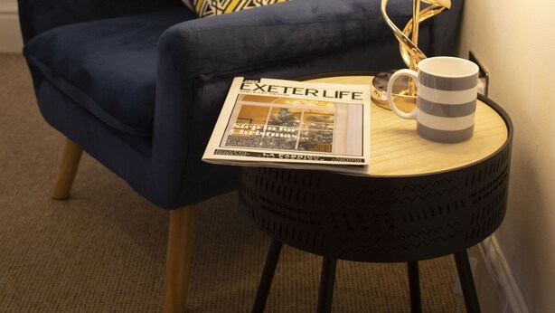 Ett mysigt rum med en blå fåtölj, en mönstrad prydnadskudde, en lampa med en spiralformad fot, en mugg och en bok med titeln ”EXETER LIFE”.