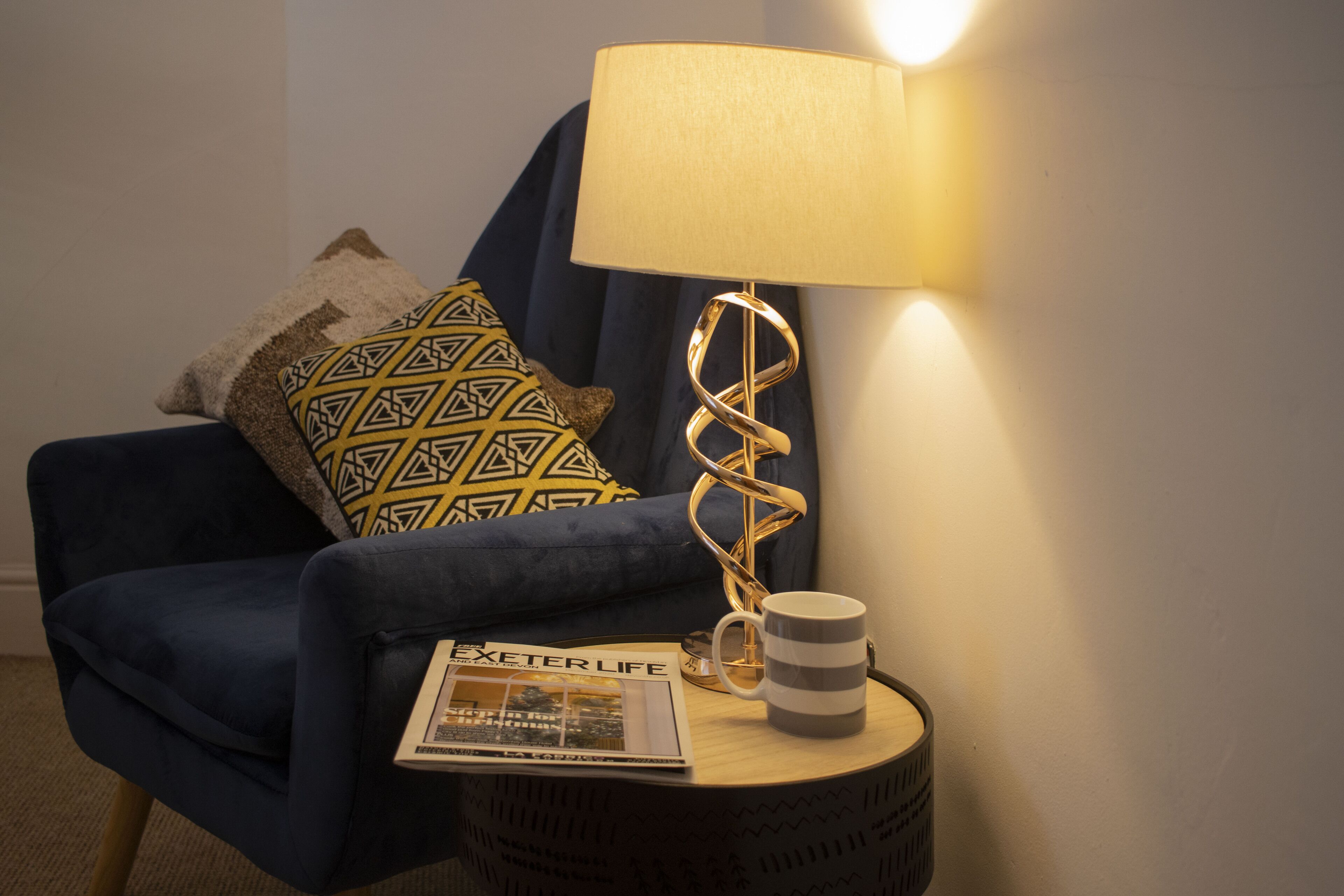 Un fauteuil bleu avec des coussins à motifs, une lampe au design en spirale, une tasse à rayures et un magazine posés sur une petite table ronde.