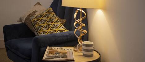 Un fauteuil bleu avec des coussins à motifs, une lampe au design en spirale, une tasse à rayures et un magazine posés sur une petite table ronde.