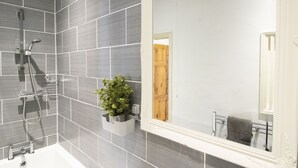 Een badkamer met grijze betegelde muren, een wit ligbad, een grote spiegel en een houten deur.