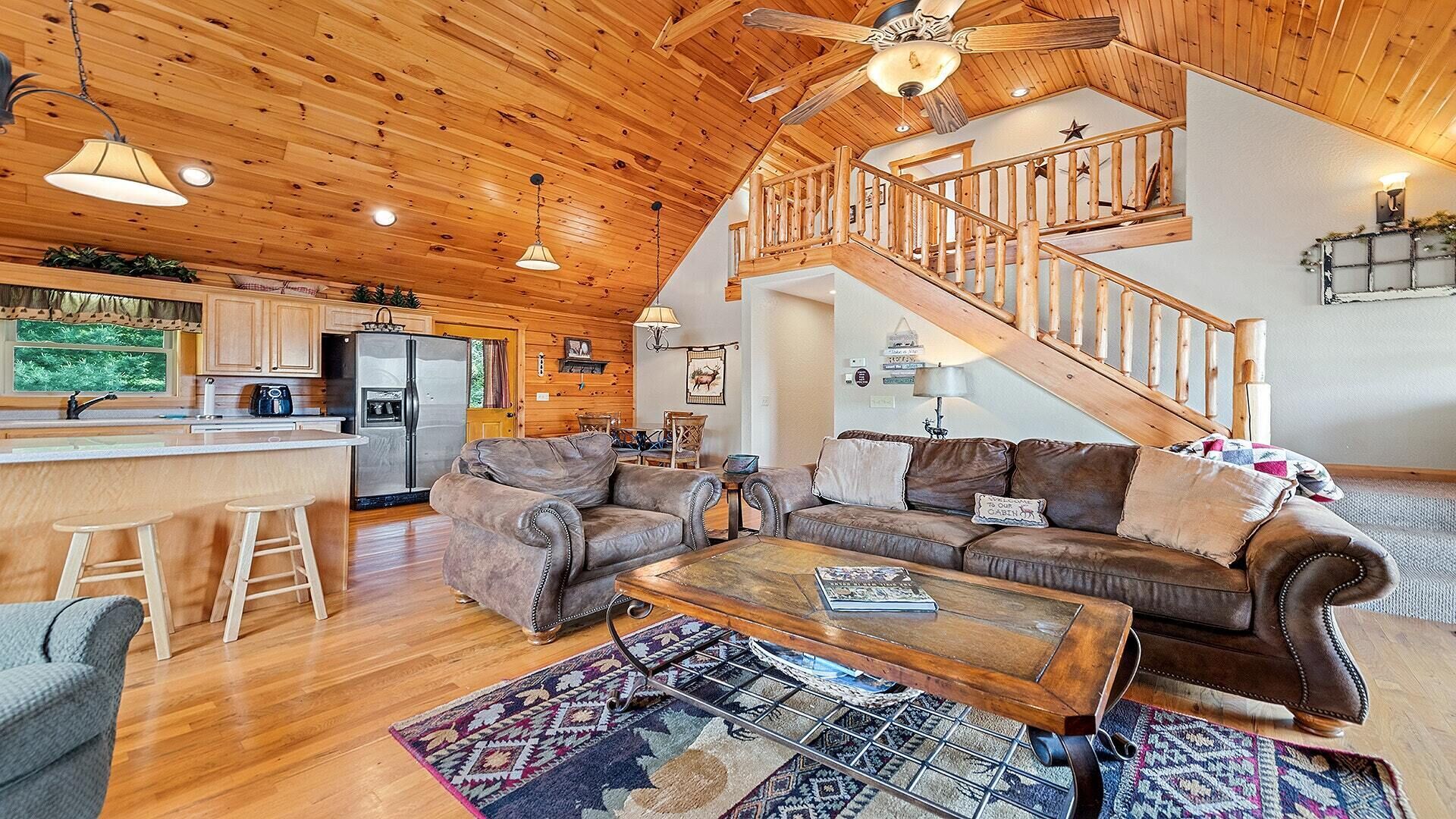 A spacious living area with a wooden ceiling, a central seating area with leather sofas, a coffee table, and a staircase leading to an upper level.
