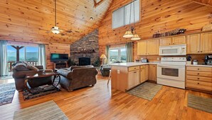 A cozy cabin interior with a stone fireplace, wooden floors, a dining area, and a kitchen with wooden cabinets and appliances. - Serenity Hill 42 Midland Drive: 3  BR, 3  BA Cabin / Bungalow in Waynesville, Sleeps 6 (Waynesville)