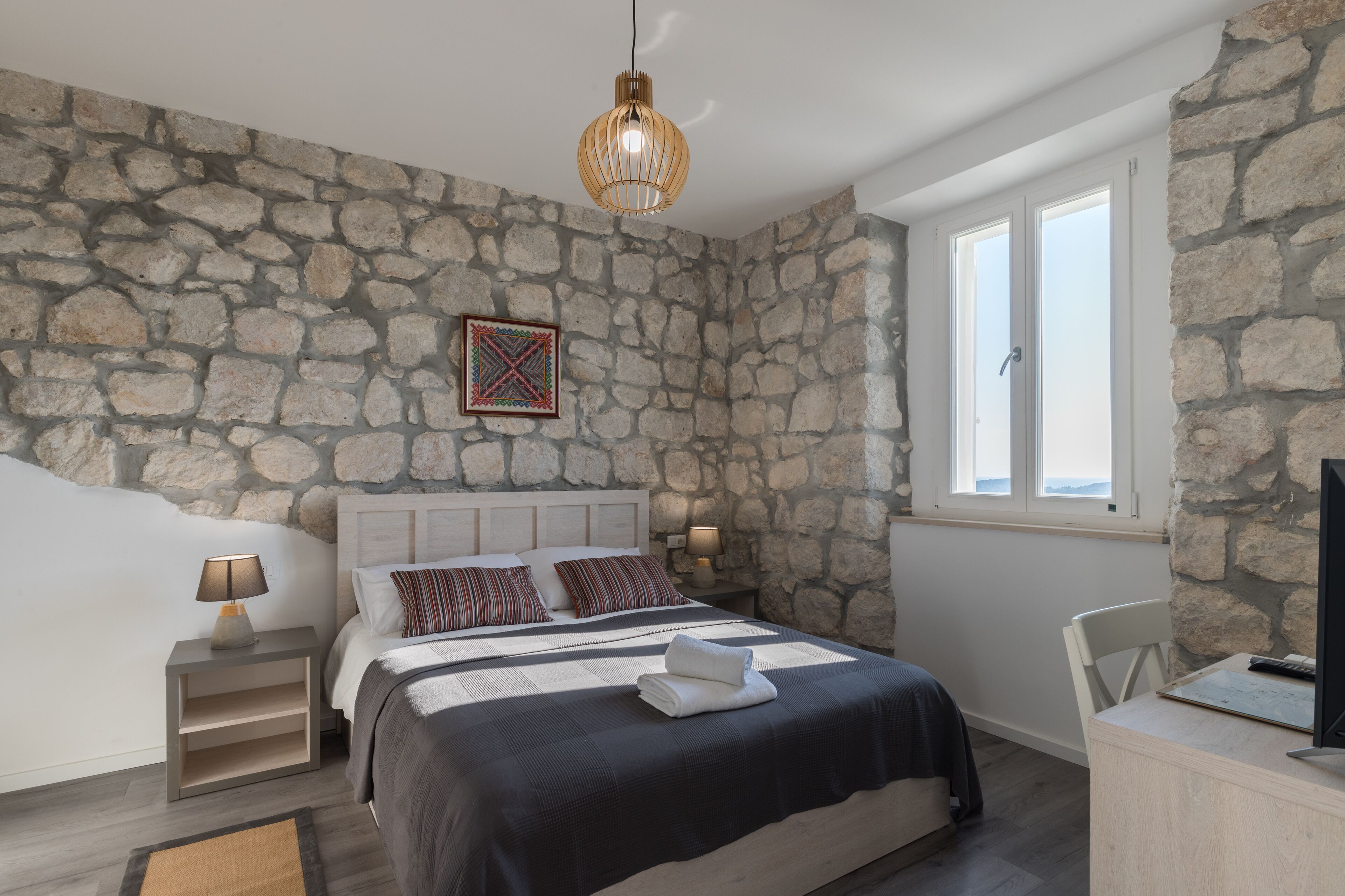 A bedroom with a stone wall, a bed with a black and white bedspread, a bedside table with a lamp, and a window with a view.