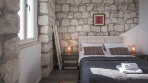 Un dormitorio con pared de piedra, cama con almohadas, mesita de noche con lámpara y ventana.