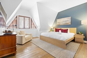 Ein modernes Schlafzimmer mit einem großen Bett, einem Schreibtisch und einem Stuhl. Es gibt Fenster mit Jalousien, ein Bild an der Wand und eine Lampe auf dem Nachttisch.