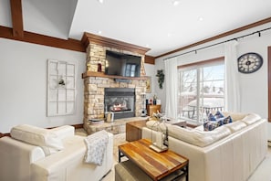 A living room with a stone fireplace, white sofas, a wooden coffee table, and a large window with a view of a snowy deck. - Le Plateau Ski In/Out w/ 3bdrs w/ Pool Access (Mont-Tremblant)
