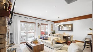 Ett rymligt vardagsrum med en vägg av sten, ett stort fönster, ett soffbord i trä, en beige soffa och en matplats med ett bord och stolar i trä.