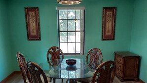 Un comedor con una mesa de vidrio rodeada por cuatro sillas, una araña y dos cuadros en las paredes.