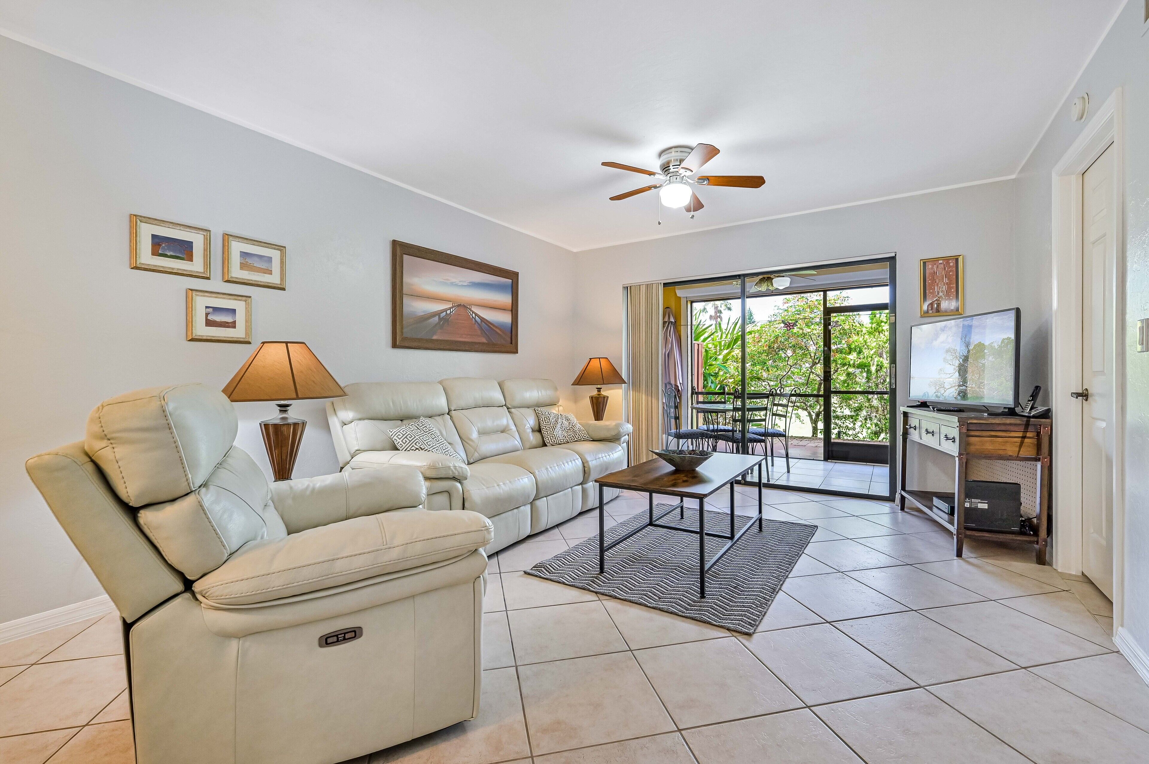 A spacious living room with a beige leather sofa, a coffee table, and a ceiling fan. There are framed pictures on the wall and a TV on a wooden stand.