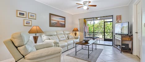 A spacious living room with a beige leather sofa, a coffee table, and a ceiling fan. There are framed pictures on the wall and a TV on a wooden stand.