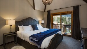 Une chambre avec un grand lit, deux tables de chevet, un lustre et un balcon offrant une vue sur les arbres et les bâtiments.