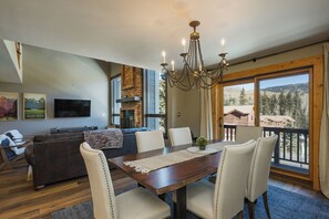 A dining area with a wooden table, six chairs, and a chandelier. There is a fireplace, a television, and a couch in the background.