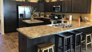 A modern kitchen with dark cabinetry, granite countertops, a stainless steel refrigerator, a microwave, and a gas stove. There are three bar stools and a vase with greenery on the island.