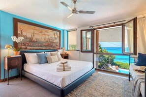 A bedroom with a bed, a ceiling fan, a nightstand, a lamp, a painting, and a view of the ocean.