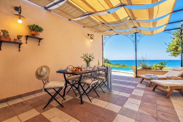 Una terraza con una mesa, sillas, un sombrero, plantas en macetas y vista al mar.