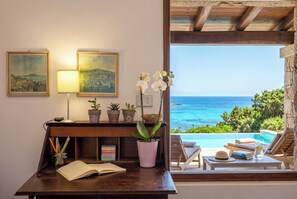 A desk with a book, a lamp, and potted plants, overlooking a pool with lounge chairs and a view of the sea.