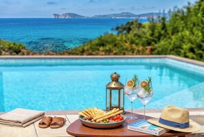 Un entorno junto a la piscina con vistas al mar, una bandeja con aperitivos, una bebida y un sombrero.