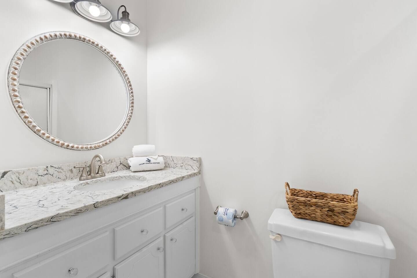 A bathroom with a marble sink, a round mirror with bead trim, a white toilet, and a wicker basket on the toilet tank.