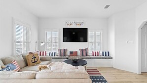 A living room with a beige sofa, a coffee table, and a TV mounted on the wall. There are striped pillows and a rug with an American flag pattern.
